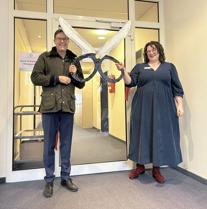 Paul Bomke, Geschäftsführer, und Alessa Boebel, fachliche Leiterin, mit einer symbolischen übergroßen Schere aus Pappe, nachdem sie ein rotes Band an der Tür durchtrennt und damit die Schule offiziell eröffnet haben.