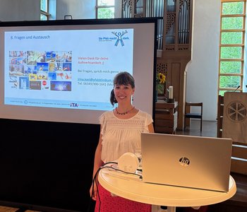 Irina Kast bei einem Vortrag mit Laptop im Vordergrund und einer Powerpointpräsentation im Hintergrund.