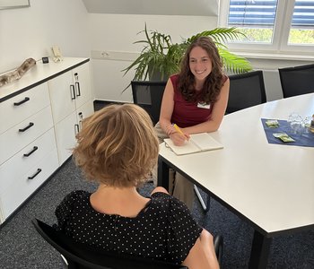 Lara Heß, eine junge Frau mit langen dunklen Haaren, sitzt mit einem Notizbuch am Tisch und berät eine Klientin, die vor ihr sitzt.