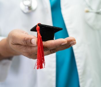 Symbolbild: Mediziner im weißen Kittel hält Mini-Doktorandenhut in der Hand 