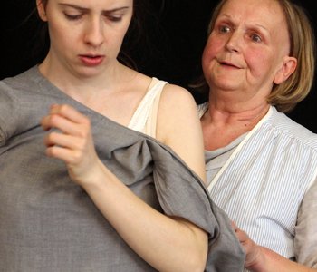 Eine ältere Frau steht hinter einer jüngeren Frau und hält sie am Arm- Szene aus dem Theaterstück "Rosa B. - beinahe vergessen"