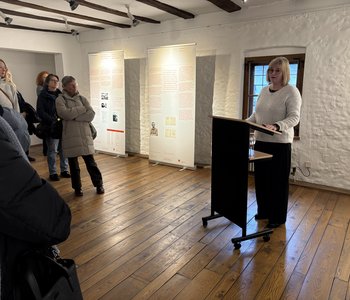 Rita Becker-Scharwatz am Rednerpult, hinter ihr zwei Infotafeln der Ausstellung, links daneben etliche Besucherinnen