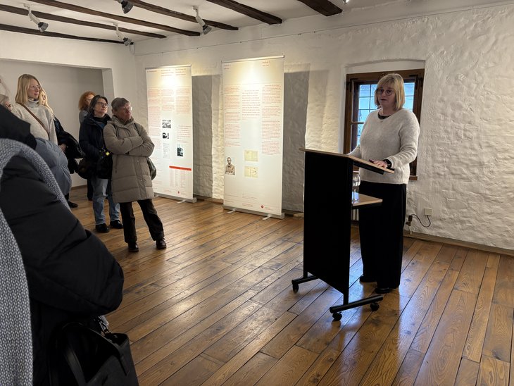 Rita Becker-Scharwatz am Rednerpult, hinter ihr zwei Infotafeln der Ausstellung, links daneben etliche Besucherinnen