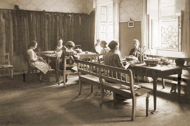 Das Bild zeigt Frauen, die an Tischen sitzen und Körbe flechten.