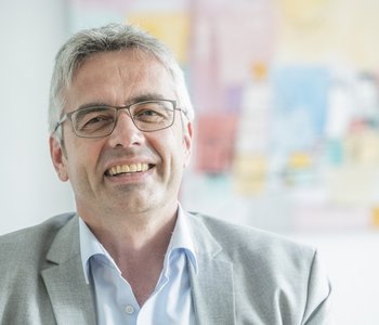 Das Bild zeigt Professor Markus Steffens. Er hat graues Haar, trägt einen grauen Anzug und eine Brille. Im Hintergrund ist ein buntes Bild an der Wand zu sehen.