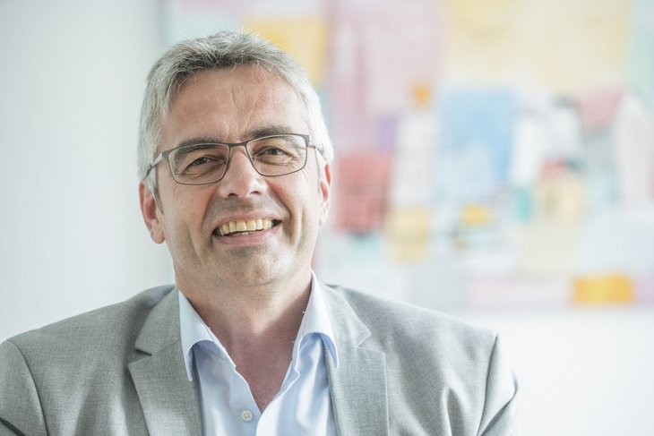 Das Bild zeigt Professor Markus Steffens. Er hat graues Haar, trägt einen grauen Anzug und eine Brille. Im Hintergrund ist ein buntes Bild an der Wand zu sehen.
